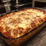 A close-up of a golden-brown Baked Ravioli Casserole topped with melted cheese and herbs in a glass baking dish.