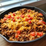 A close-up of a hearty Beef and Rice Skillet topped with melted cheddar cheese and diced tomatoes.