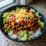 A close-up of a Beef Burrito Bowl featuring rice, seasoned ground beef, black beans, corn, tomatoes, shredded cheese, and lettuce.