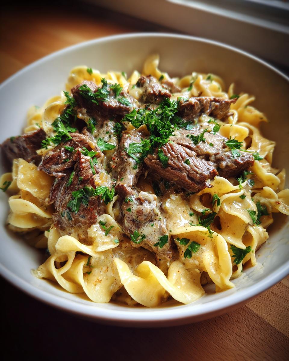 A close-up view of a bowl of Beef Stroganoff With Egg Noodles, topped with sliced beef and fresh parsley.