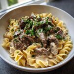 A close-up of Beef Stroganoff With Egg Noodles in a white bowl, topped with fresh parsley.