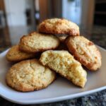 A close-up of a stack of golden-brown cake mix cookies on a white plate, with one cookie broken in half to show its soft interior.