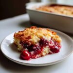 A slice of cherry Dump Cake on a white plate, showcasing a golden crust and juicy cherry filling.