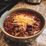 A close-up of a bowl filled with hearty Chili Con Carne, topped with shredded cheese and kidney beans.