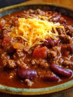 A close-up of a bowl filled with hearty Chili Con Carne, topped with shredded cheddar cheese.
