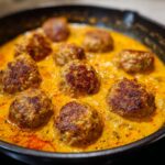 Close-up of delicious Creamy Coconut Curry Meatballs simmering in a rich, orange sauce in a cast-iron skillet.