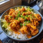 A close-up of a Crispy Bang Bang Chicken Bowl, featuring fried chicken pieces drizzled with sauce over rice and shredded lettuce, garnished with green onions and sesame seeds.