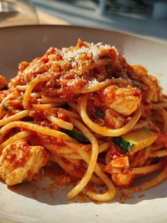 A close-up of a bowl of Italian chicken pasta with chunks of chicken, zucchini slices, and a rich tomato sauce, topped with grated cheese.