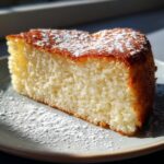A close-up of a slice of Italian Ricotta Cake, dusted with powdered sugar, on a light blue plate.