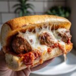 A hand holding a delicious meatball sub overflowing with juicy meatballs, marinara sauce, and melted cheese.
