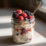A close-up of Mixed Berry Overnight Oats in a mason jar, topped with fresh raspberries, blueberries, and blackberries, with a spoon.