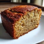 A close-up of a moist slice of banana cake on a white plate, showing its tender crumb and golden-brown crust.