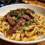A close-up of a white bowl filled with One Pot Creamy Spicy Garlic Steak Pasta, topped with sliced steak and fresh parsley.