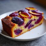 A close-up of a slice of Plum Torte Cake, showcasing juicy plums baked into a golden cake with a dusting of powdered sugar.