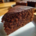 A close-up of a moist slice of rich chocolate cake on a white plate, with the crumb texture clearly visible.