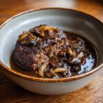 A close-up of a juicy Salisbury Steak With Gravy, topped with mushrooms and onions in a rich brown sauce.