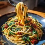 A fork lifts a swirl of Spaghetti Spinach With Sun Dried Tomatoes, showing pasta, spinach, and sun-dried tomatoes, topped with grated cheese.