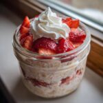Close-up of Strawberries Cream Overnight Oats in a glass jar, topped with fresh strawberries and whipped cream.