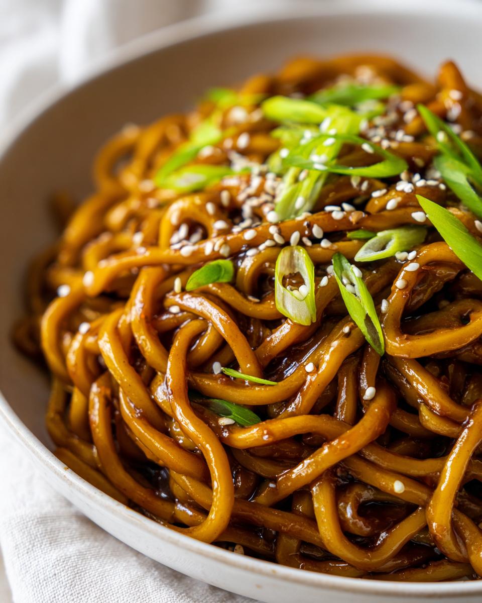 A close-up, aesthetic shot of glossy brown noodles coated in sauce, topped with sliced green onions and sesame seeds.