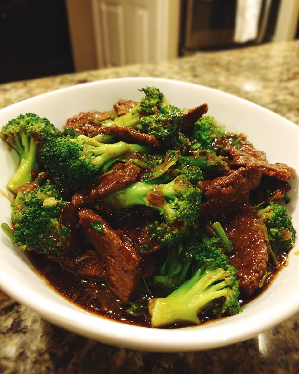 A white bowl filled with glistening Beef And Broccoli Stir Fry, featuring tender beef slices and bright green broccoli florets in a dark sauce.