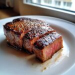Close-up of a thick, medium-rare beef steak, seasoned and sliced, resting in its juices on a white plate.