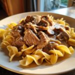 Close-up of tender beef chunks and mushrooms in creamy sauce served over egg noodles, classic Beef Stroganoff Crockpot.