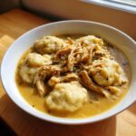 A close-up of a white bowl filled with creamy Chicken And Dumplings, featuring fluffy dumplings and shredded chicken.