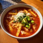 Close-up of a vibrant bowl of Chicken Tortilla Soup topped with shredded cheese, avocado, and tortilla strips.