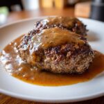 Close-up of two juicy Classic Salisbury Steak patties smothered in rich brown gravy on a white plate.