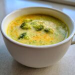Close-up of a white mug filled with creamy, bright yellow and green Broccoli Cheese Soup, featuring visible chunks of broccoli florets.