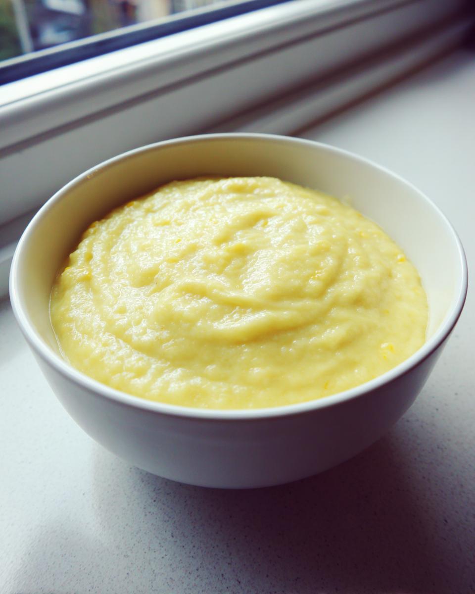A white bowl filled with thick, creamy yellow Summer Corn And Zucchini Soup, set near a window.