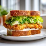 A tall, delicious Crispy Chicken Caesar Sandwich featuring golden fried chicken, romaine lettuce, and creamy dressing between two slices of toasted bread.