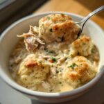 A spoonful lifting a fluffy, golden-brown dumpling and shredded chicken from a bowl of creamy Crockpot Chicken And Dumplings.