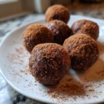 Six rich, dark date snack balls dusted heavily with cocoa powder on a white plate.