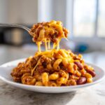 A spoonful of Easy One Pot Chili Macaroni lifted from a plate, showing melted cheese stretching between the spoon and the main dish.