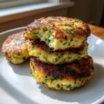 A stack of three golden brown, pan-fried Zucchini Patties with a bite taken out of the top one, showing the shredded zucchini interior.