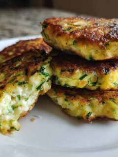 A stack of four golden-brown, pan-fried Zucchini Patties, showing the green shreds of zucchini inside the soft center.