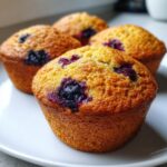 Close-up of four golden-brown Healthy Greek Yogurt Blueberry Muffins with visible juicy blueberries on a white plate.