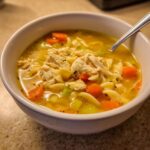 Close-up of a white bowl filled with rich, golden Chicken Noodle Soup featuring shredded chicken, carrots, celery, and egg noodles.