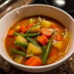 Close-up of a white bowl filled with steaming Vegetable Soup featuring chunks of carrots, potatoes, green beans, and celery in a rich broth.