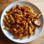 A white bowl filled with Honey Pepper Chicken Pasta featuring penne noodles coated in a glossy, dark sauce with visible chili flakes.