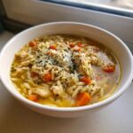 A close-up of a white bowl filled with Italian Penicillin Soup, featuring shredded chicken, orzo pasta, carrots, and topped with grated Parmesan cheese and herbs.