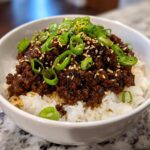A close-up of a white bowl filled with white rice topped generously with savory Korean Ground Beef Bowl and garnished with sliced green onions and sesame seeds.