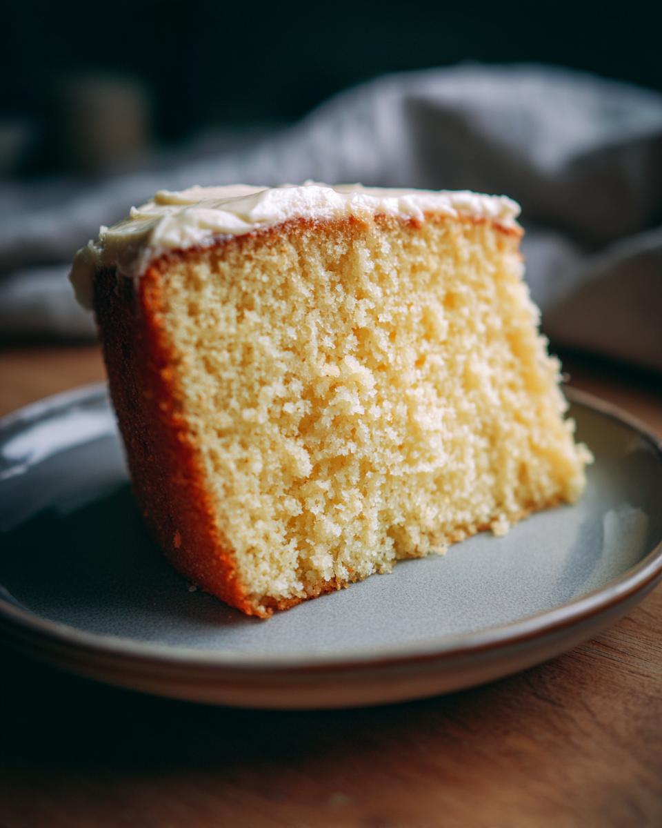 A close-up of a moist slice of homemade Vanilla Cake topped with white frosting on a small plate.