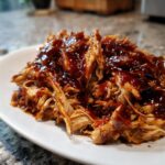 Close-up of juicy, shredded Bbq Chicken Crockpot coated in thick, glossy barbecue sauce on a white plate.