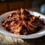 A close-up, appetizing shot of shredded Instant Pot Beef Barbacoa piled high on a white plate, glistening with sauce.