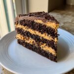 A tall slice of rich Peanut Butter Chocolate Cake showing three layers of dark chocolate cake separated by thick peanut butter frosting.