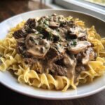 A bowl of Slow Cooker Beef Stroganoff featuring tender beef and mushrooms in a creamy sauce served over wide egg noodles, garnished with parsley.