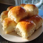 A pile of golden brown, shiny Soft Homemade Dinner Rolls stacked on a white plate, showing their fluffy interior.