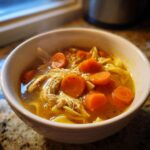 Close-up of a steaming white bowl filled with rich chicken noodle Crockpot Soup, featuring shredded chicken and bright orange carrot slices.
